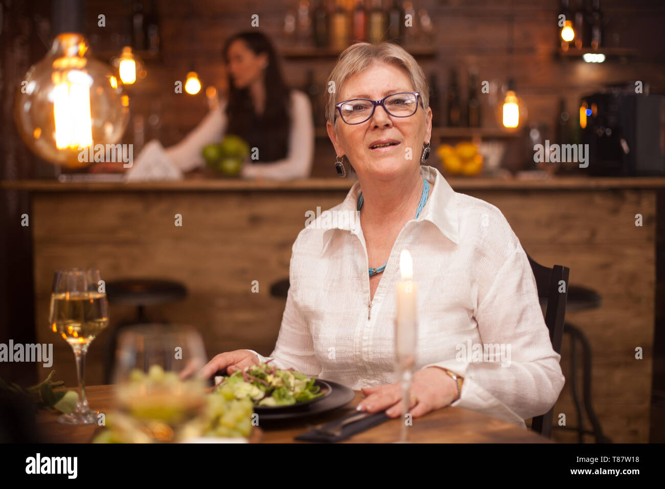 Portrait de jolie vieille dame regardant la caméra tout en appréciant son repas dans un restaurant. Un verre de vin blanc. Salade de légumes. Banque D'Images
