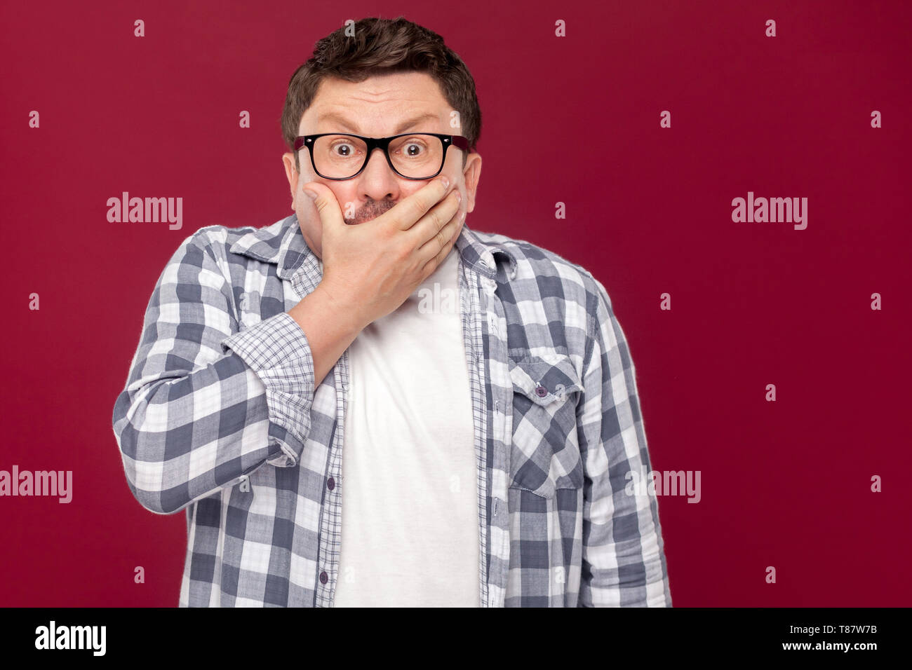 Portrait of middle aged man in casual chemise à carreaux et des lunettes debout, couvrant sa bouche et looking at camera avec peur f Banque D'Images