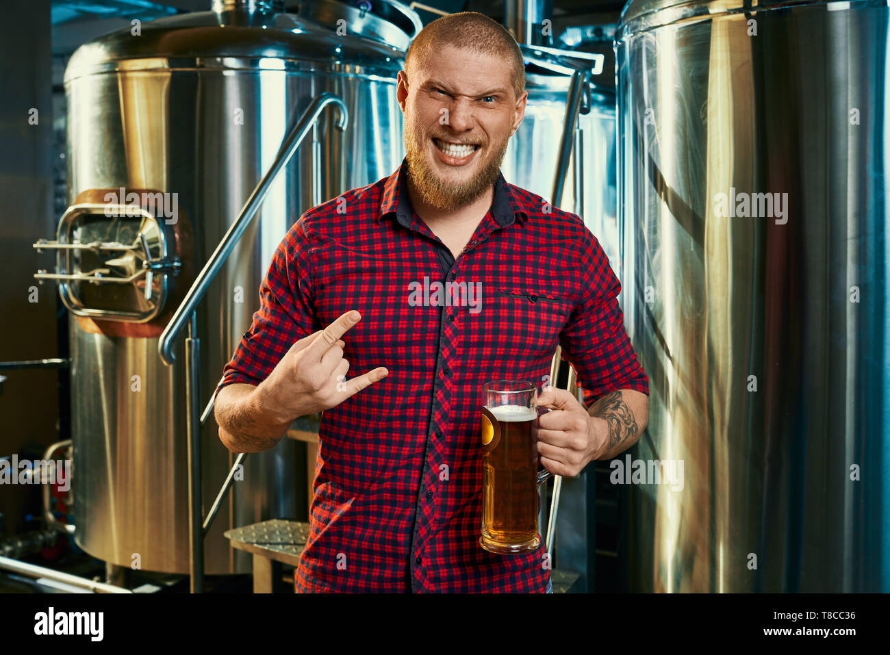 Vue avant fo fou hipster barbu debout dans la brasserie, maintien de la bière et montrant signe de rock. Young male looking at camera et posant avec près de verre machines de brassage. Concept de boisson. Banque D'Images