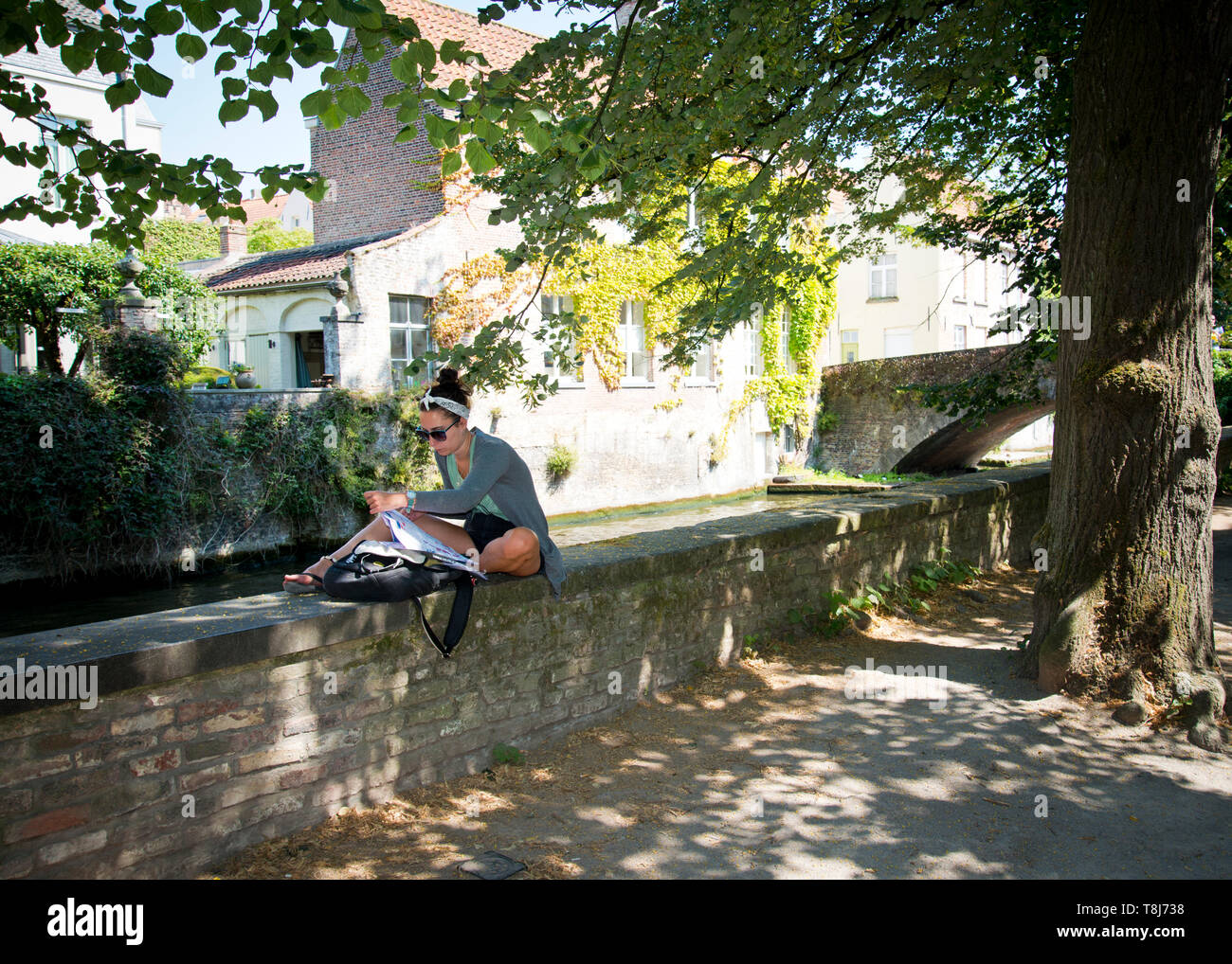 Jeune femme seule, assise sur le mur bas par le canal, regardant la carte pour les directions et le repos, Bruges, Belgique, Europe, Banque D'Images
