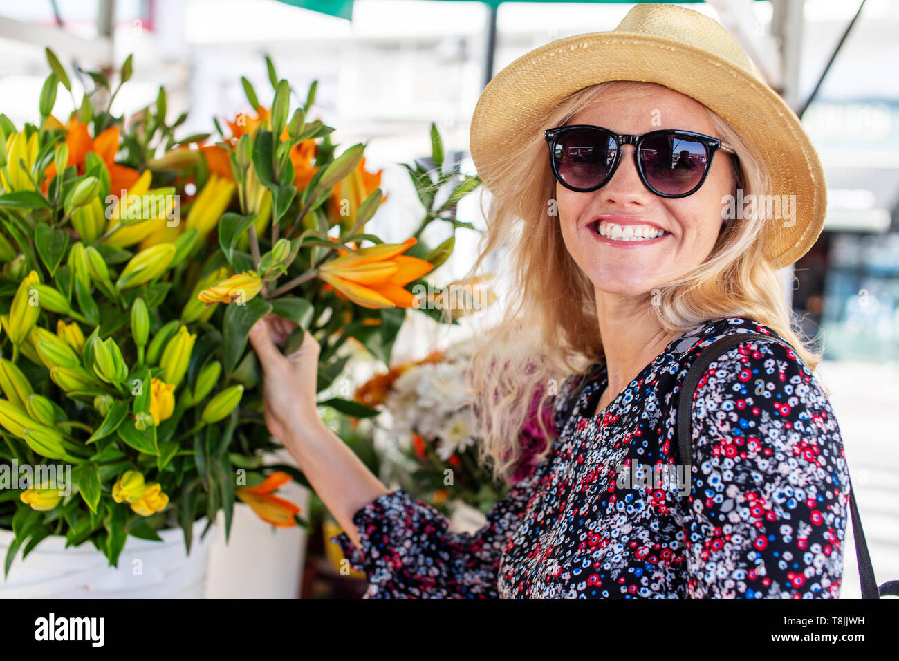 Blonde femme méditerranéenne en lunettes de soleil et chapeau de toucher des fleurs au marché intérieur Banque D'Images