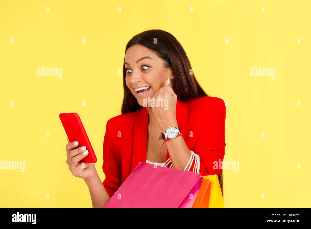 Euphorique excité Mid adult woman using smartphone holding shopping bags poing de pompage dans le bonheur isolé sur fond jaune. Ethnique multiculturelle Banque D'Images