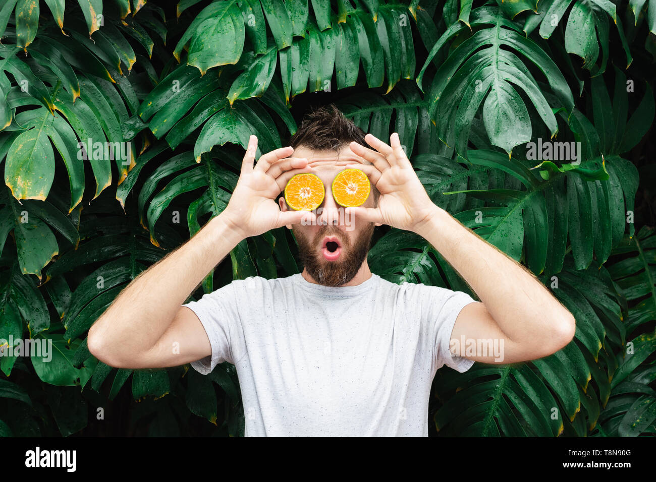 Jeune homme barbu tenant tranches de mandarine orange devant ses yeux, surpris sur son visage. Arrière-plan de feuilles tropicales, de copier l'espace. Banque D'Images