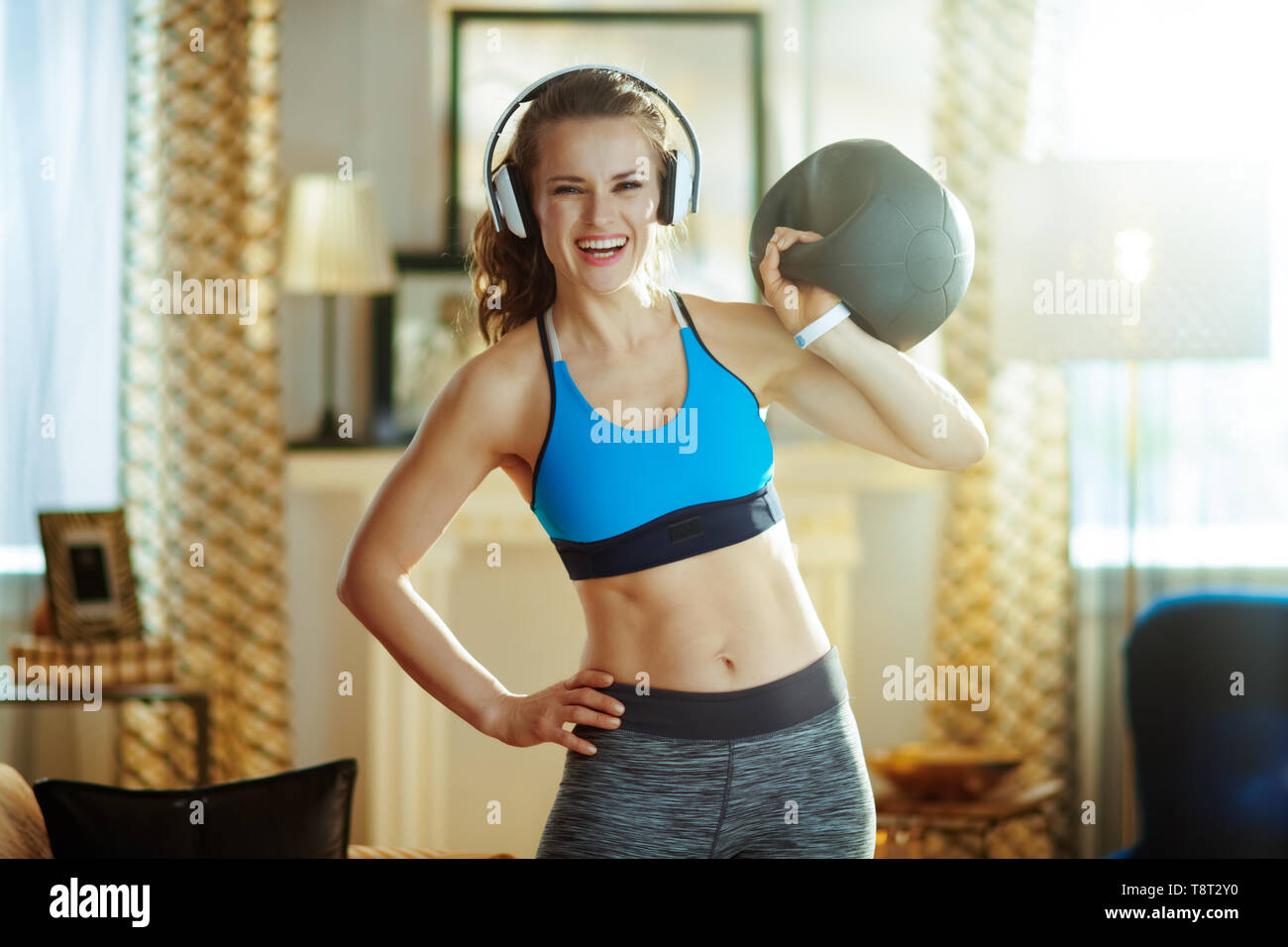 Happy active femme dans les écouteurs en vêtements de sport avec équipement d'entraînement fonctionnel dans la maison moderne. Banque D'Images