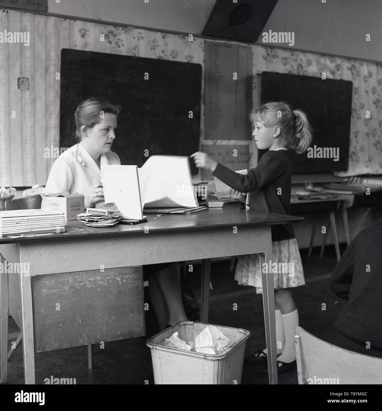 Années 1960, écolière historique parlant à son professeur qui est assis à son bureau dans la salle de classe, Angleterre, Royaume-Uni. Banque D'Images