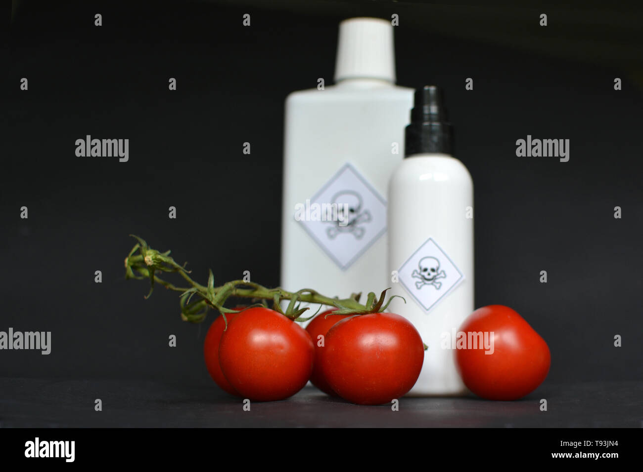 Pesticides dans les produits alimentaires agricoles avec des tomates rouges en premier plan et bouteilles blanches floues avec un poison de l'étiquette d'avertissement sur fond noir Banque D'Images