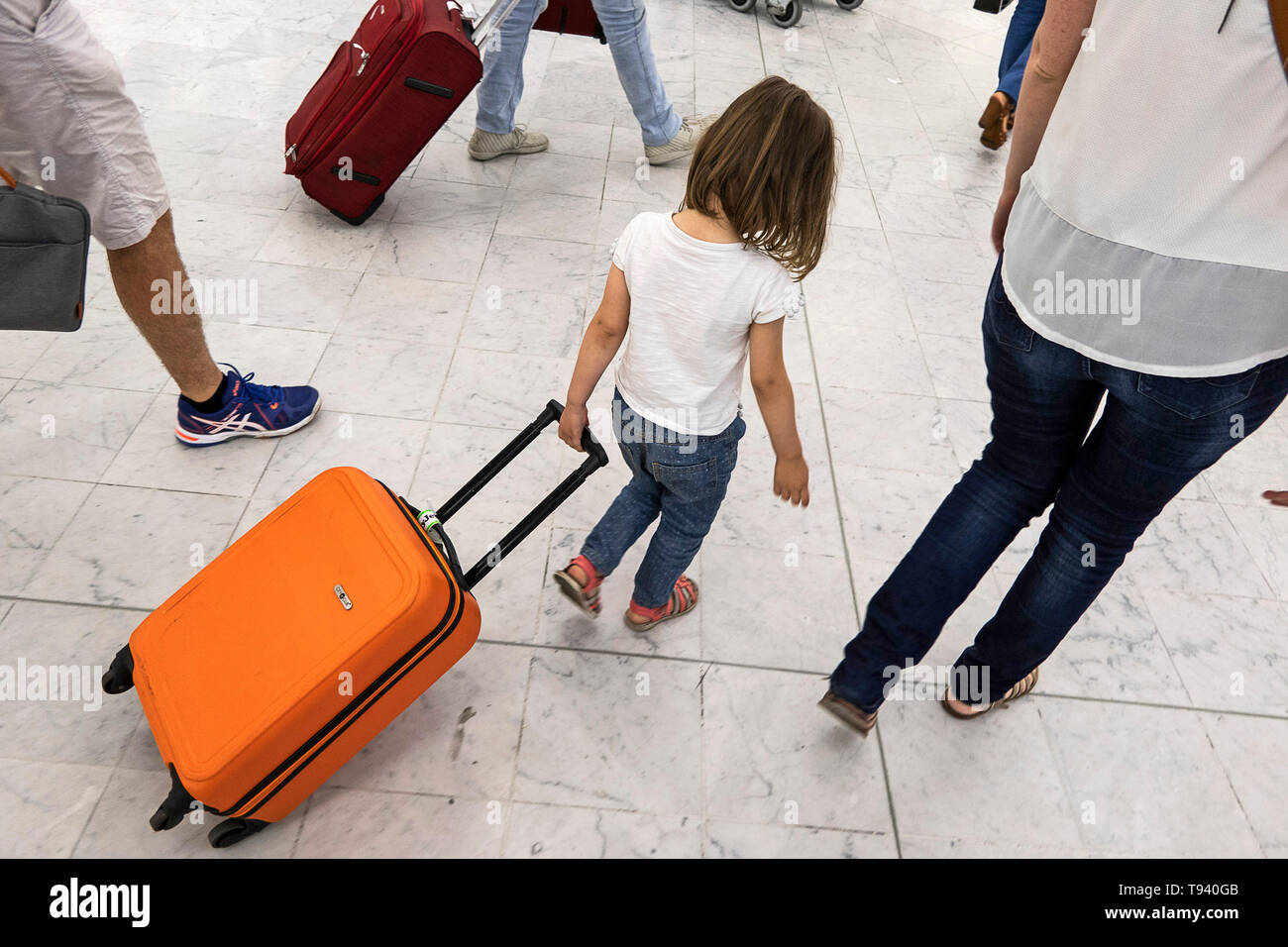 Petite fille, la tête en bas, tirant une valise trolley orange dans un hall de l'aéroport d'Orly Banque D'Images