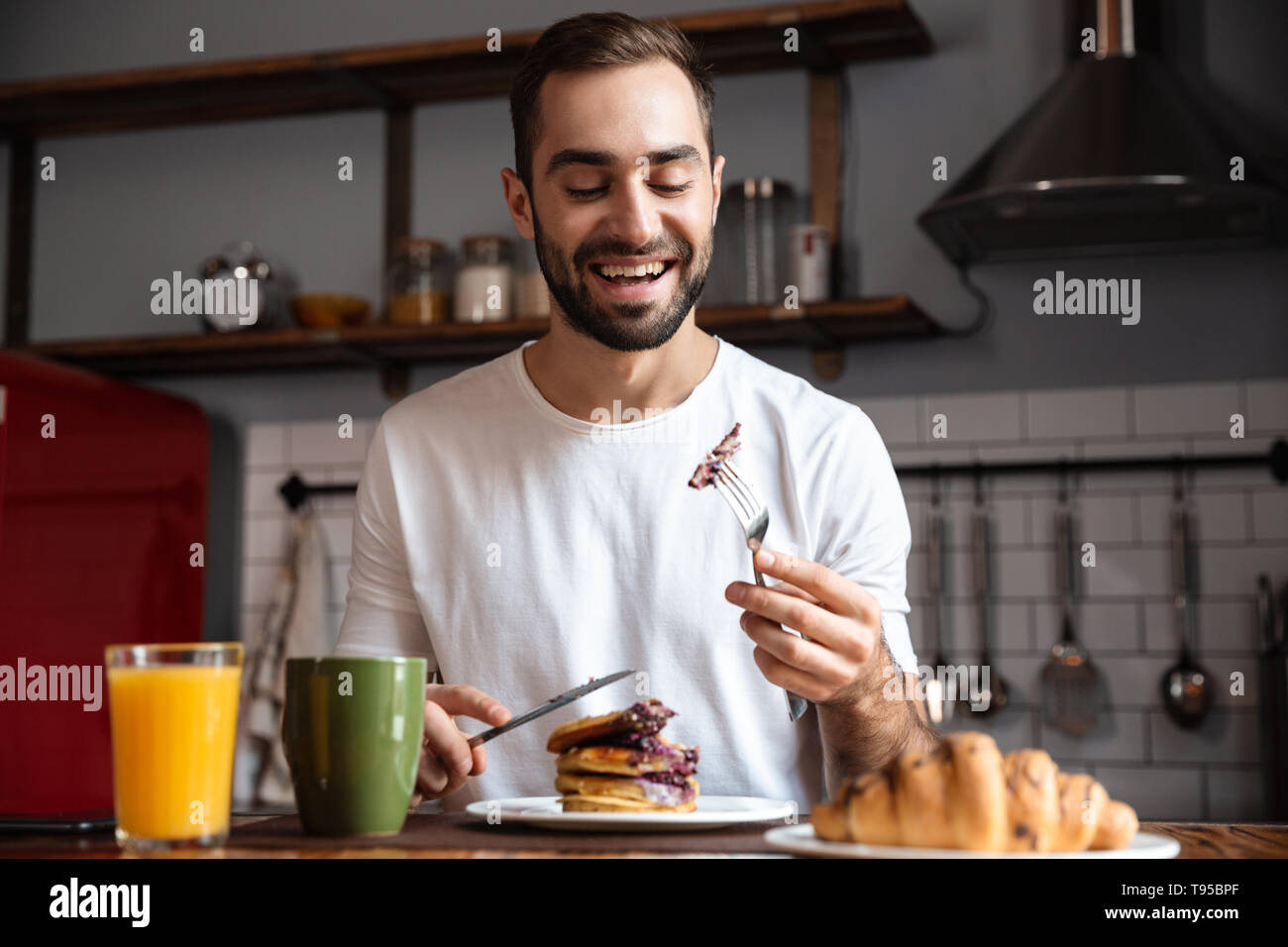 Image de brunette guy 30s assis à table et de manger des aliments pendant le petit-déjeuner à appartement moderne Banque D'Images