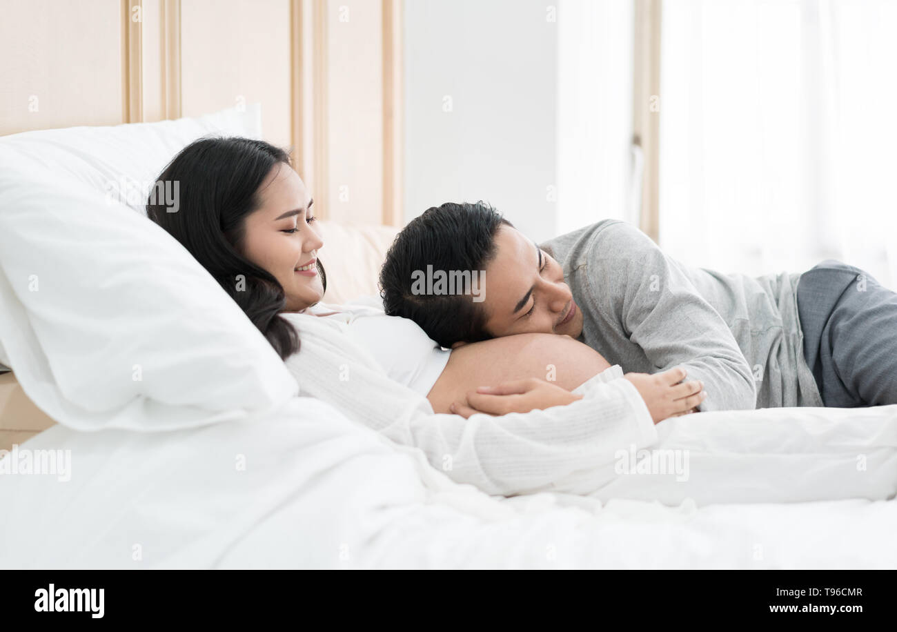 Bel homme asiatique est à l'écoute de sa belle épouse asiatique enceinte le ventre et sourit. Famille amour concept. Banque D'Images