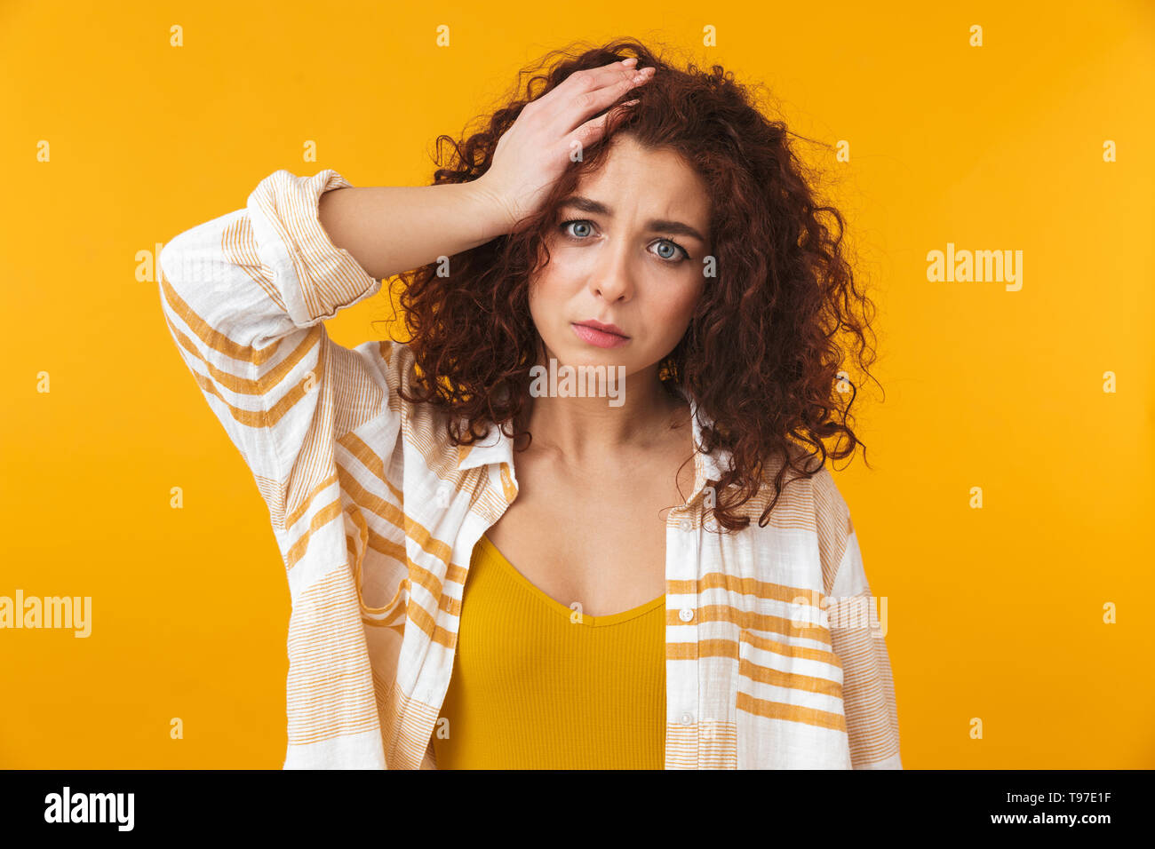 Image de femme perplexe 20s avec des cheveux bouclés saisissant sa tête, isolé sur fond jaune Banque D'Images