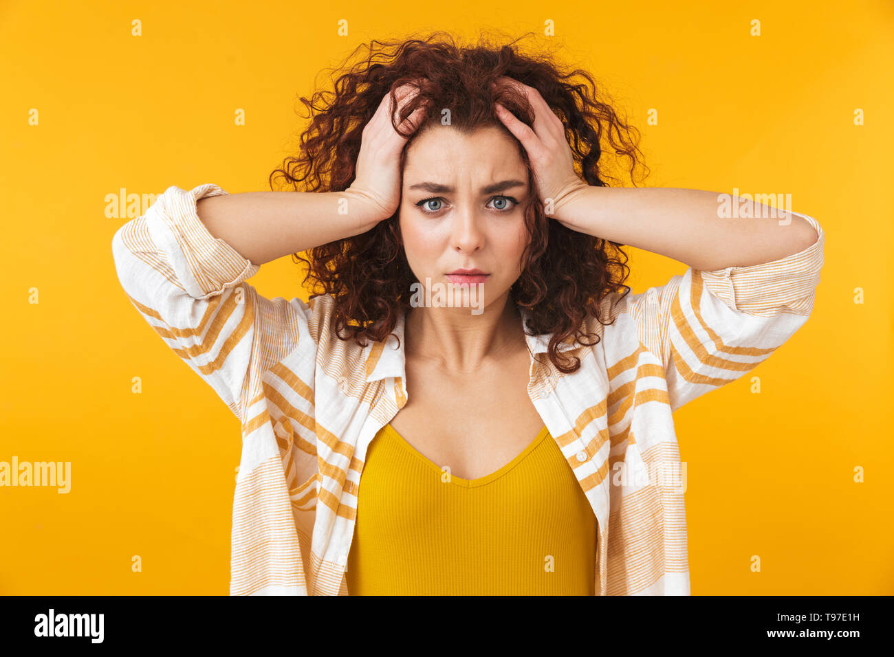 Image de femme tendue 20s avec des cheveux bouclés saisissant sa tête, isolé sur fond jaune Banque D'Images