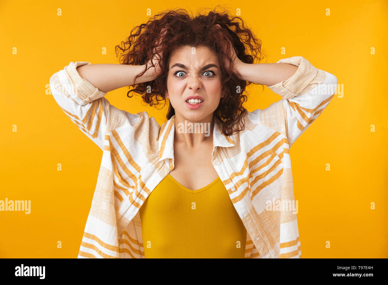 Image de femme agressive 20s avec des cheveux bouclés saisissant sa tête, isolé sur fond jaune Banque D'Images