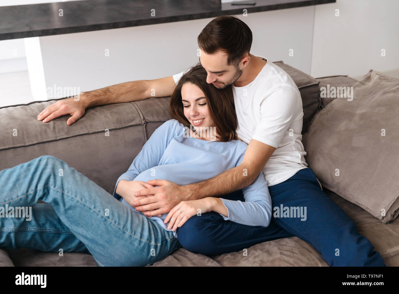 Image de portrait femme enceinte et bel homme dans des vêtements décontractés hugging ensemble à la maison tout en étant allongé sur le canapé dans la salle de séjour Banque D'Images