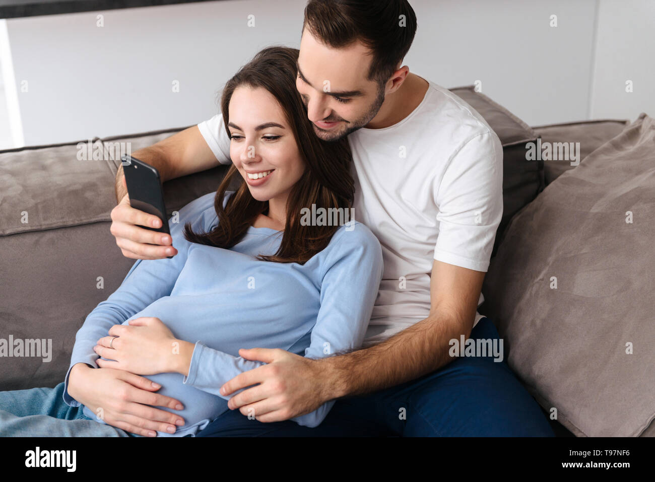 Image de femmes enceintes séropositives et bel homme en vêtements de détente allongé sur un canapé dans la salle de séjour à la maison et using smartphone Banque D'Images