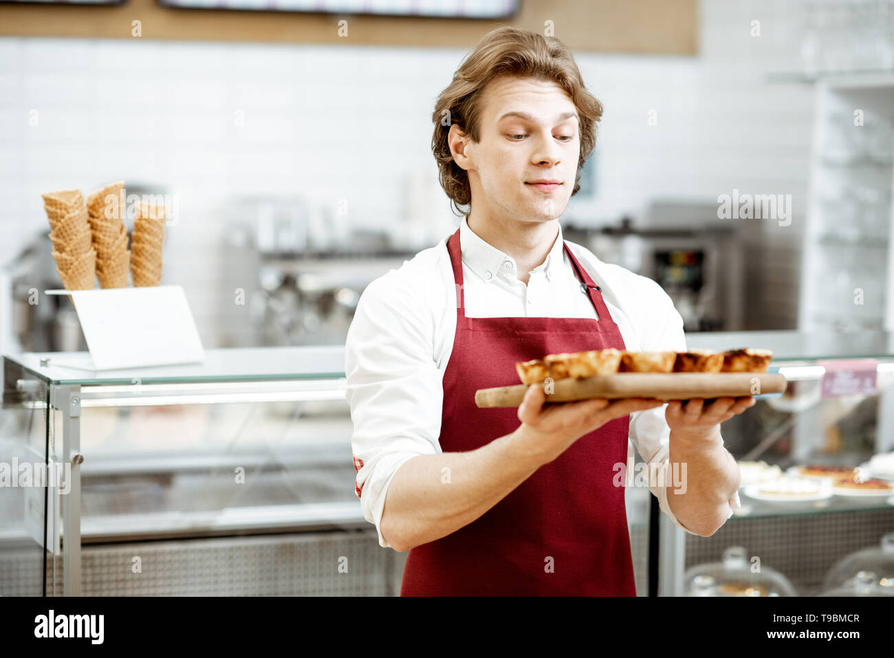 Portrait of a handsome vendeur ou pâtissier en rouge tablier, debout avec des petits pastel de nata dans la pâtisserie Banque D'Images