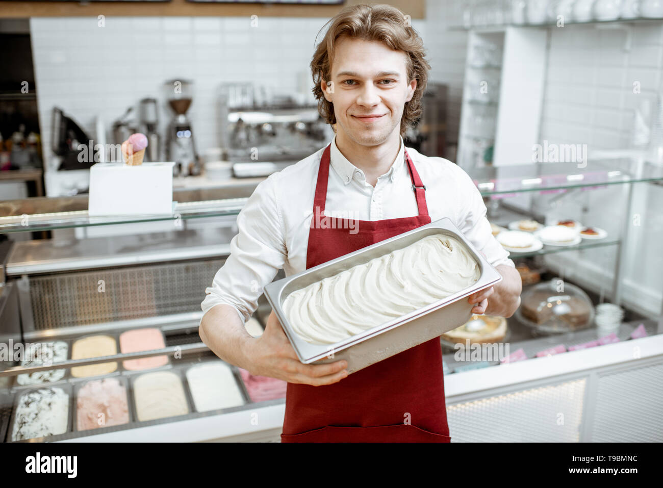 Portrait of a handsome vendeur ou pâtissier en tablier rouge holding tray with ice cream dans la boutique Banque D'Images