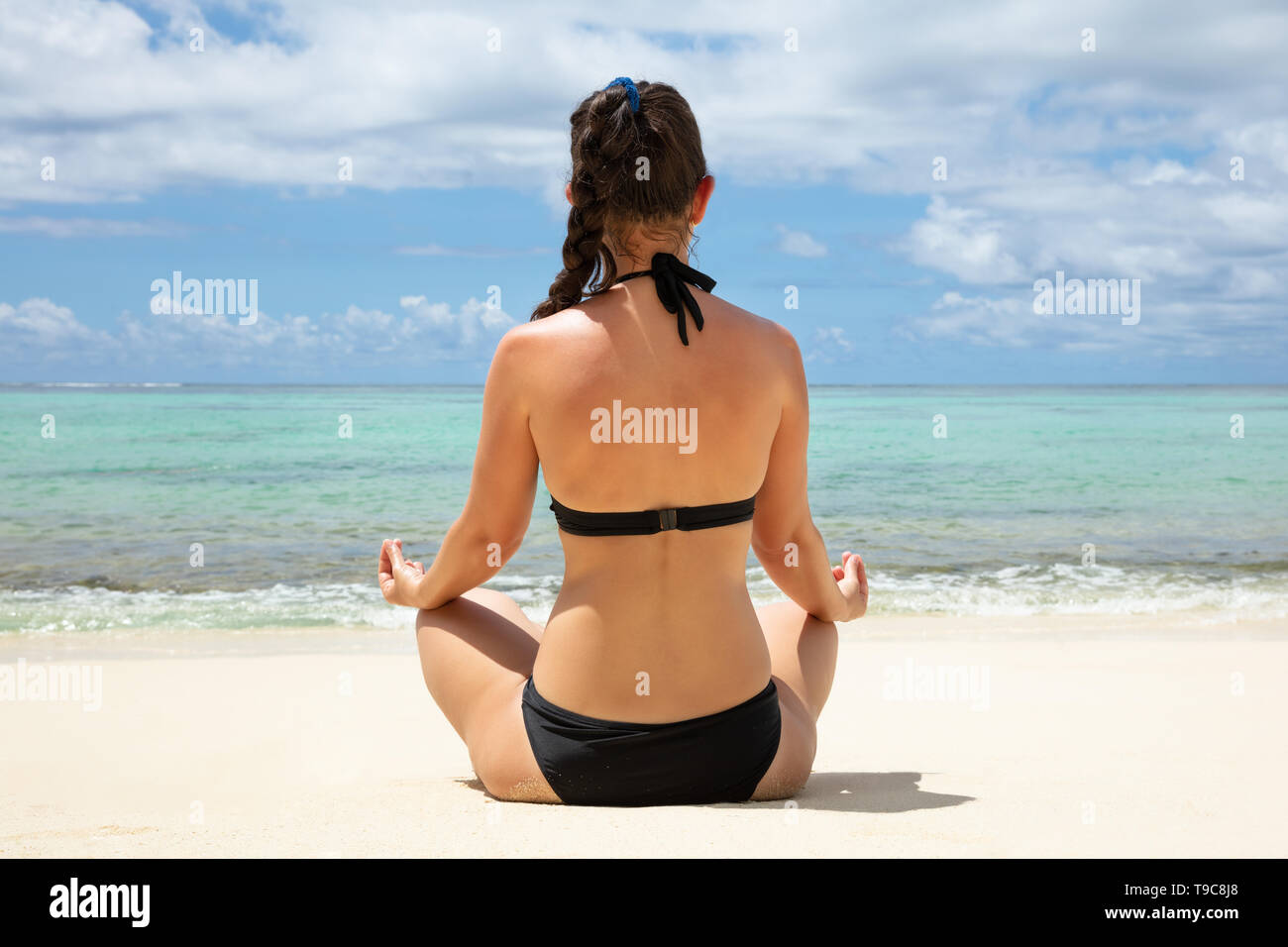 Vue arrière d'une femme en bikini noir méditant sur plage près de la mer turquoise Banque D'Images