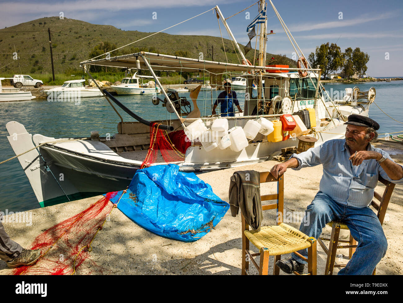 Pêcheur grec prendre pause cigarette assis sur une chaise avec bateau de pêche traditionnel derrière et vue sur le port et au-delà Banque D'Images
