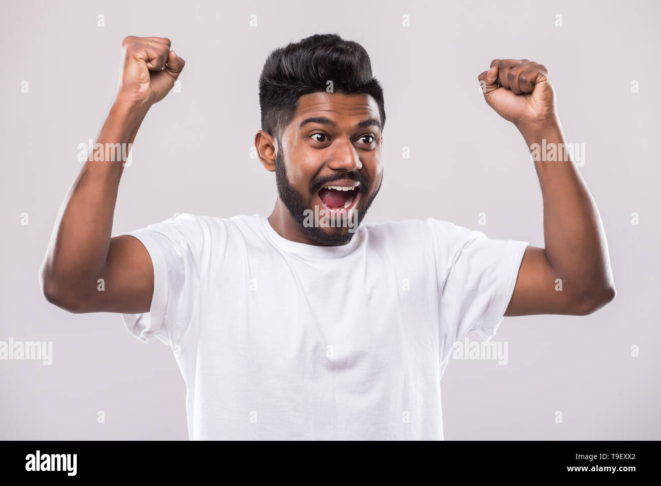 J'ai gagné. Happy young Indian man gesturing et smiling while standing against white background Banque D'Images