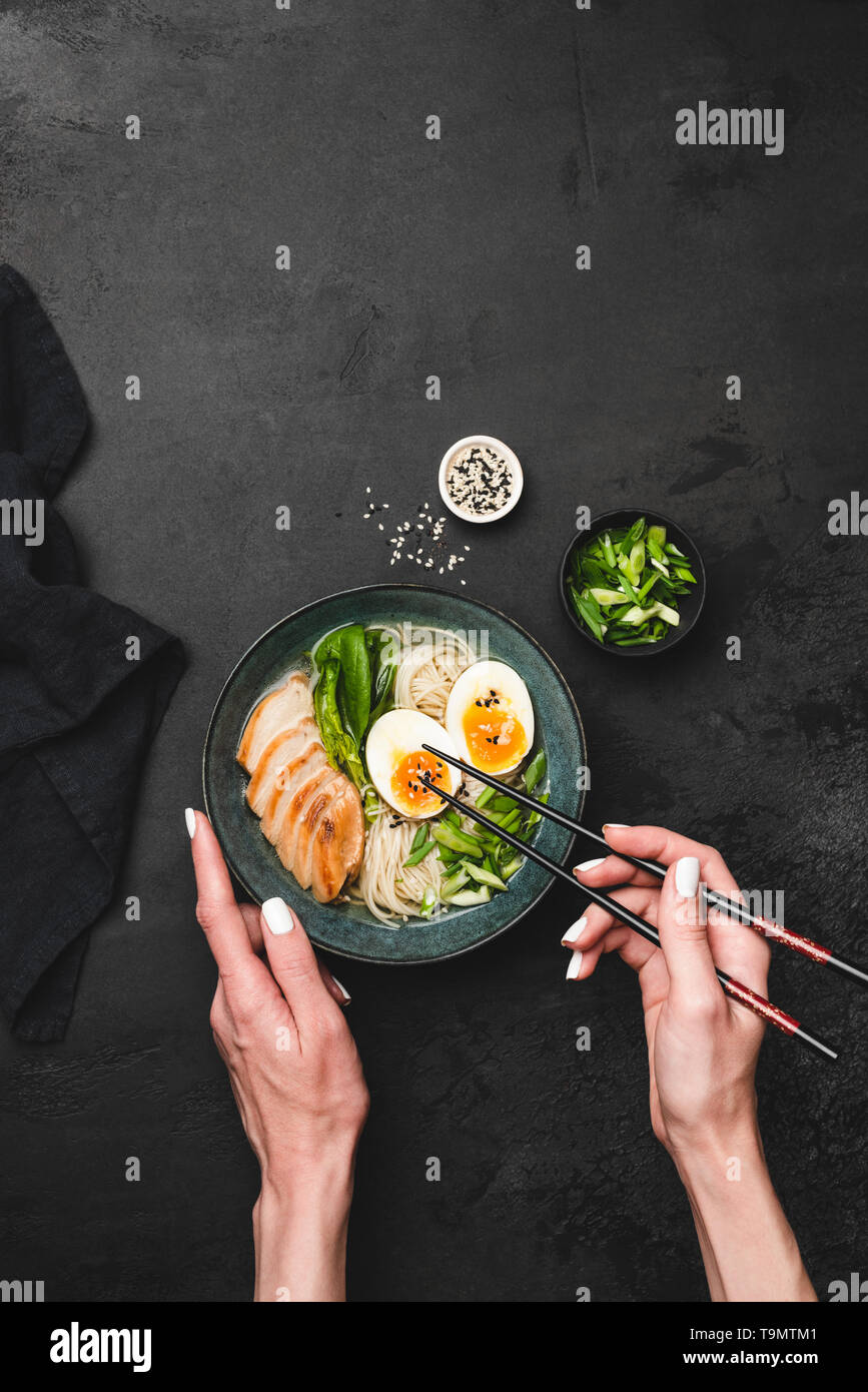 Manger la soupe de Ramen avec du poulet et des oeufs. Mains tenant au-dessus des baguettes bol de ramen japonais bol. Arrière-plan de béton noir, vue de dessus de table Banque D'Images