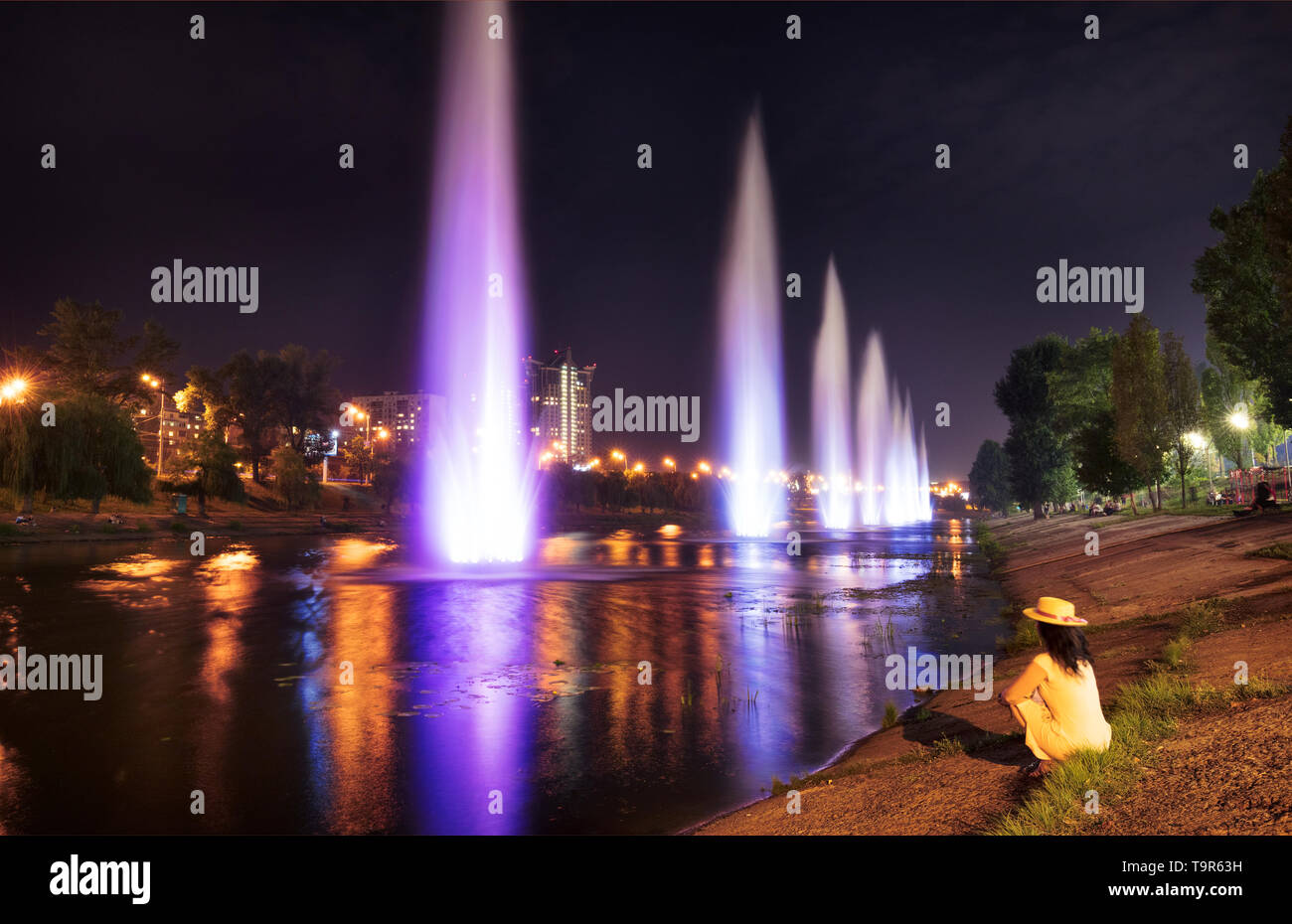 Une fille dans une robe jaune est situé sur la rive de la rivière, sur l'herbe et regarde les belles fontaines, lumineux et haute sur le canal d'Rusanovskaya Banque D'Images