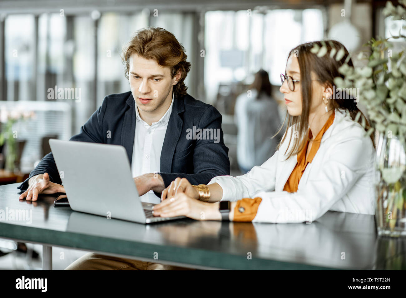 Jeune homme et femme ayant une conversation d'affaires avec ordinateur portable tout en étant assis au bar ou bureau spacieux moderne Banque D'Images