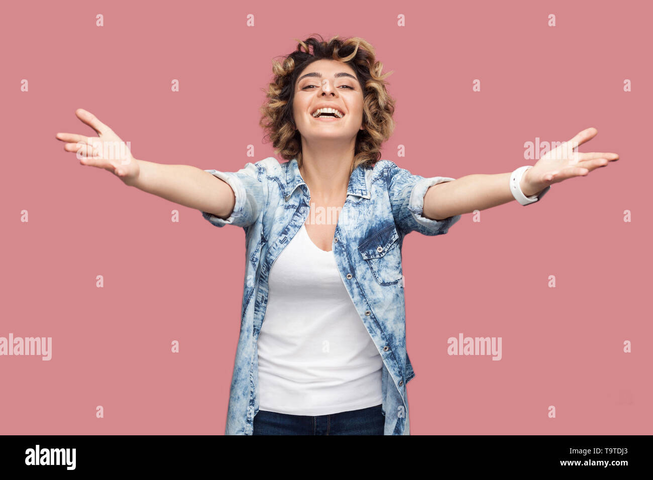 Donne-moi dans ses bras. Portrait de jeune femme avec les cheveux bouclés casual shirt bleu debout avec les bras levés pour embrasser et looking at camera, smi dentelée Banque D'Images