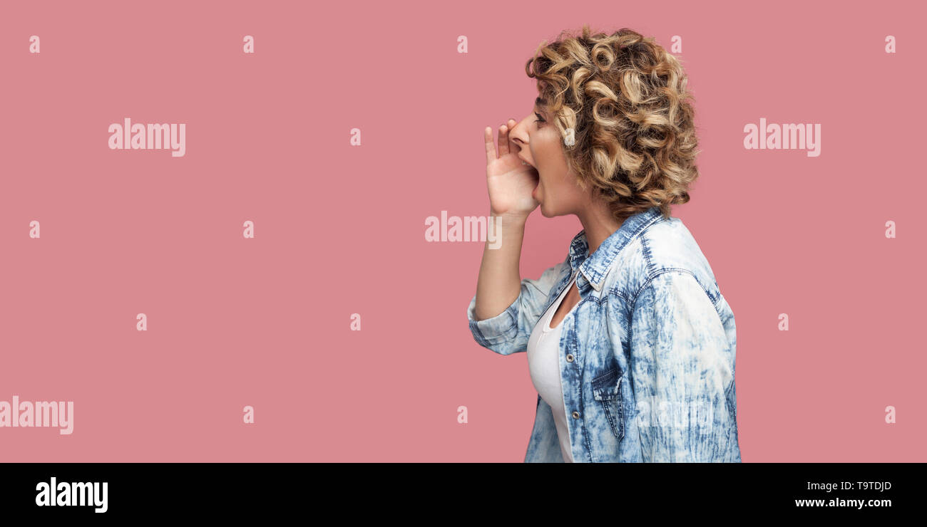Vue côté profil portrait of young woman with curly hairstyle in casual shirt bleu, debout à l'avant près de la main tenant la bouche et je cris. Banque D'Images