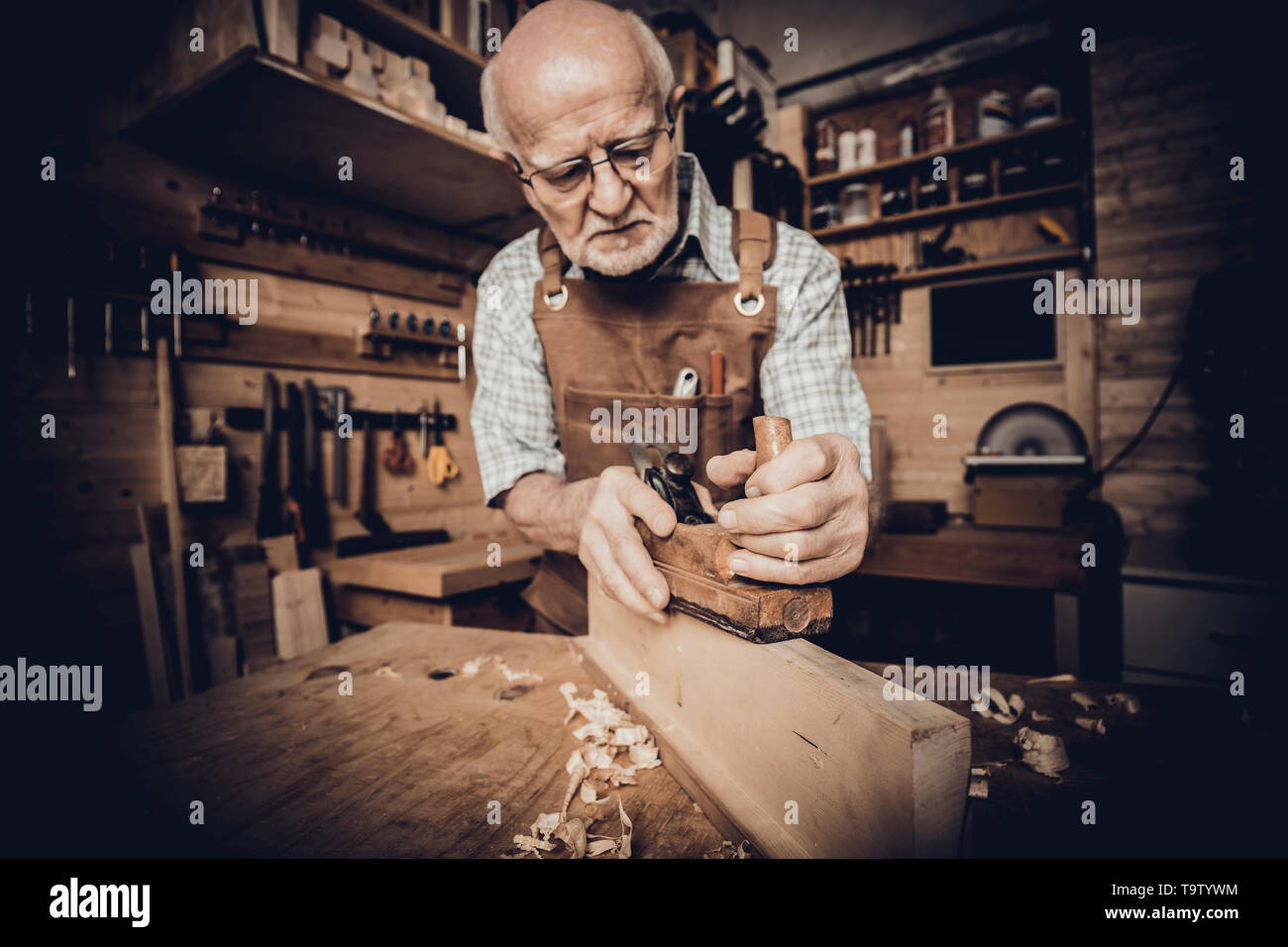 Vieux carpenter avec des lunettes adoucit un morceau de bois avec la main à l'intérieur de son atelier d'avion Banque D'Images