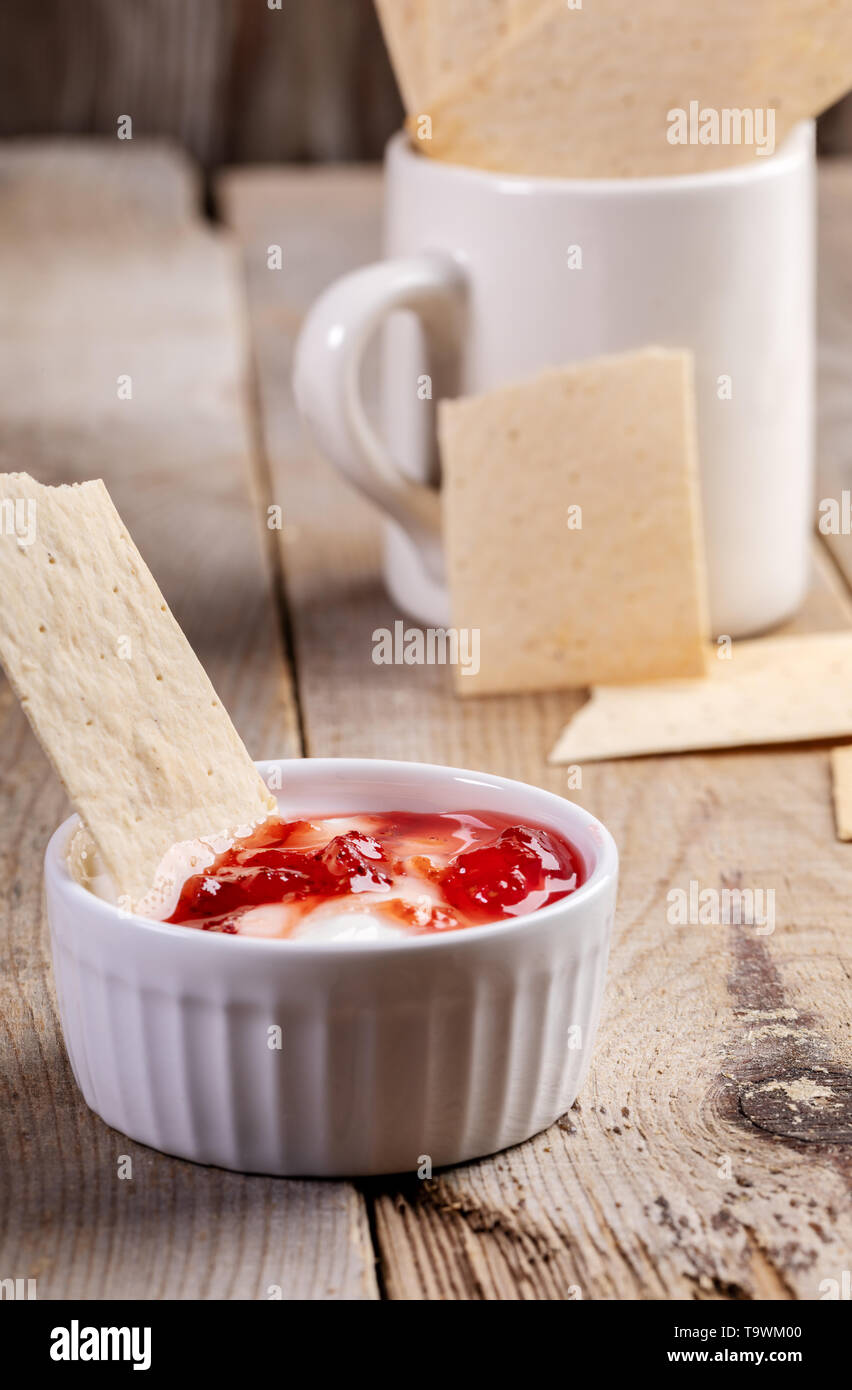 Close-up white ceramic bowl avec de la crème et de la confiture de fraise avec des biscottes de blé et blé blanc mug avec crispbreads sur la vieille table en bois rustique. Banque D'Images