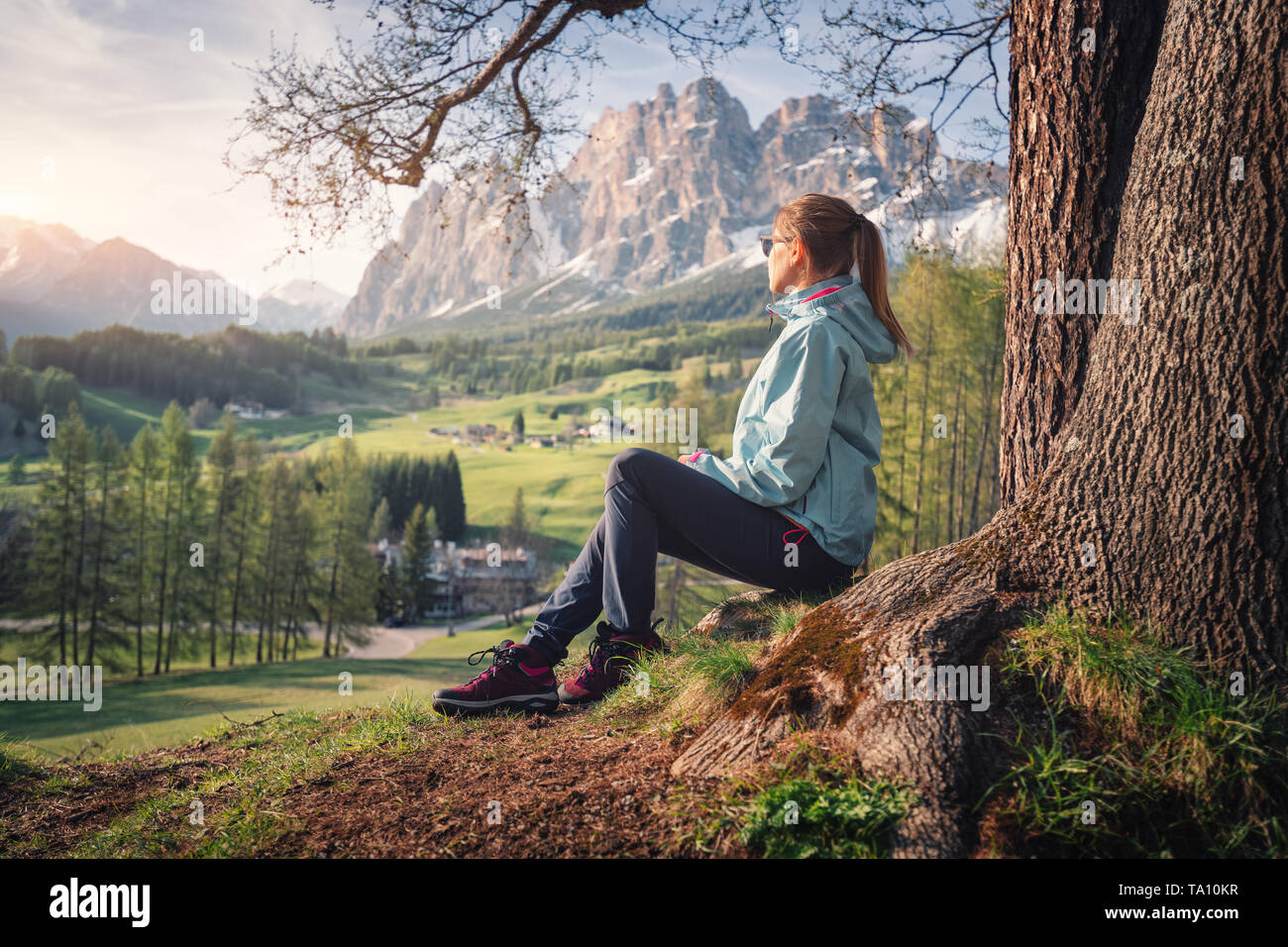 Belle jeune femme à lunettes de soleil et veste bleue est assis sur la colline sous l'arbre au coucher du soleil. Paysage printemps coloré avec fille sportive, meado Banque D'Images