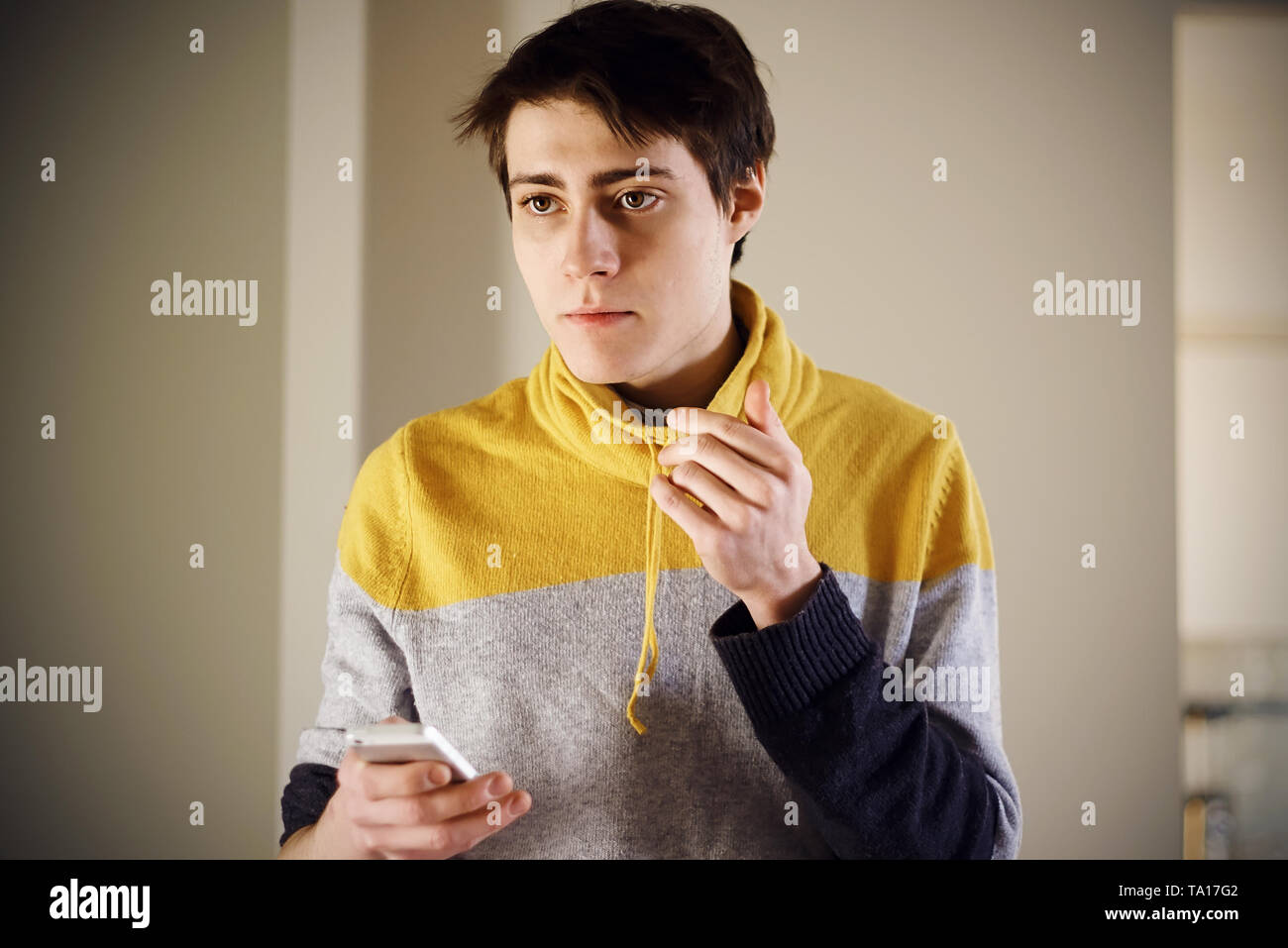 Un beau jeune gars dans un chandail jaune est titulaire d'un téléphone dans ses mains et regarde pensivement sur le côté, alors qu'éclairé par le soleil du matin Banque D'Images