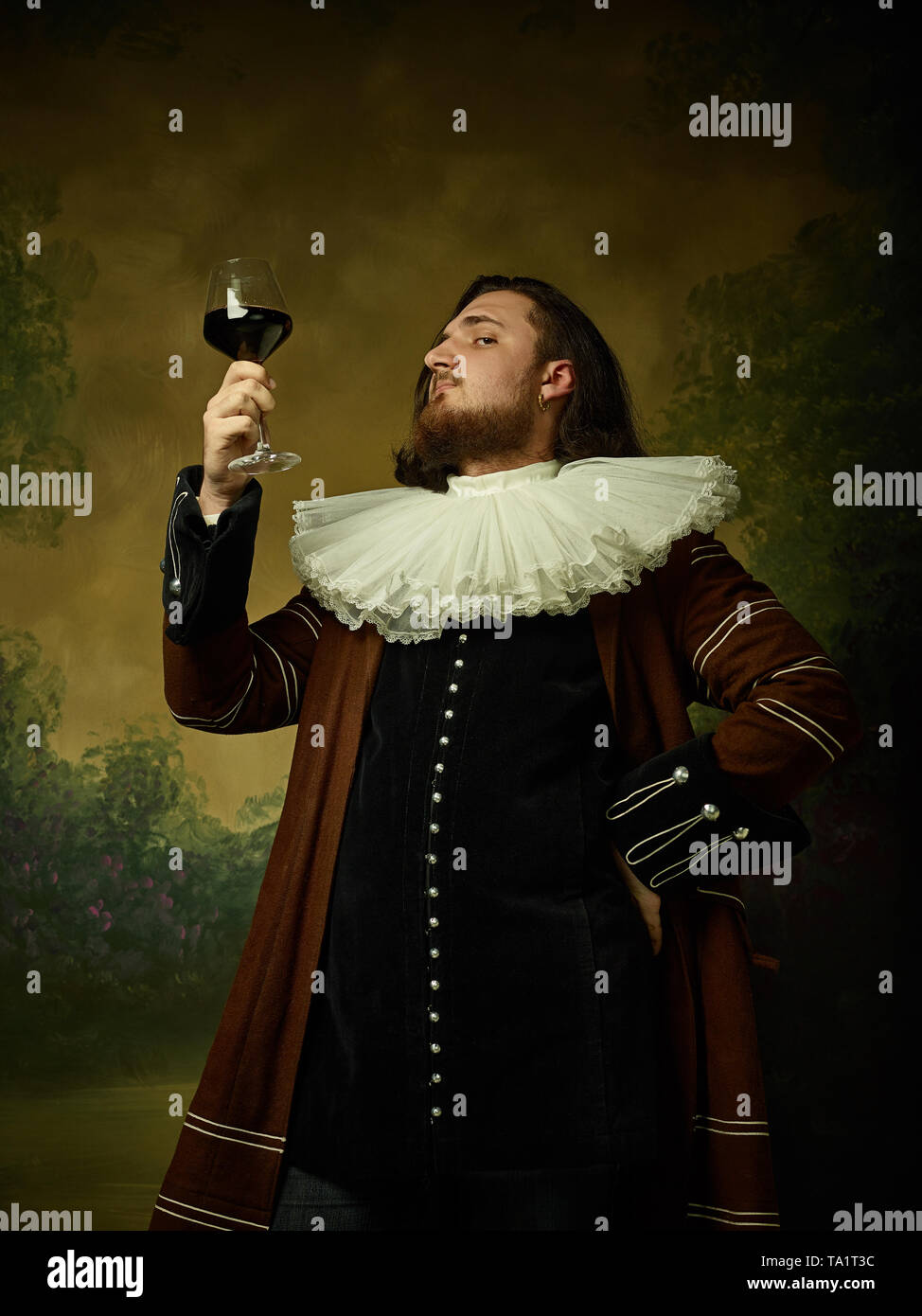 Jeune homme comme un chevalier médiéval sombre sur fond de studio. Portrait de modèle masculin en costume rétro. Tenant un verre de vin rouge. Les émotions humaines, compar Banque D'Images