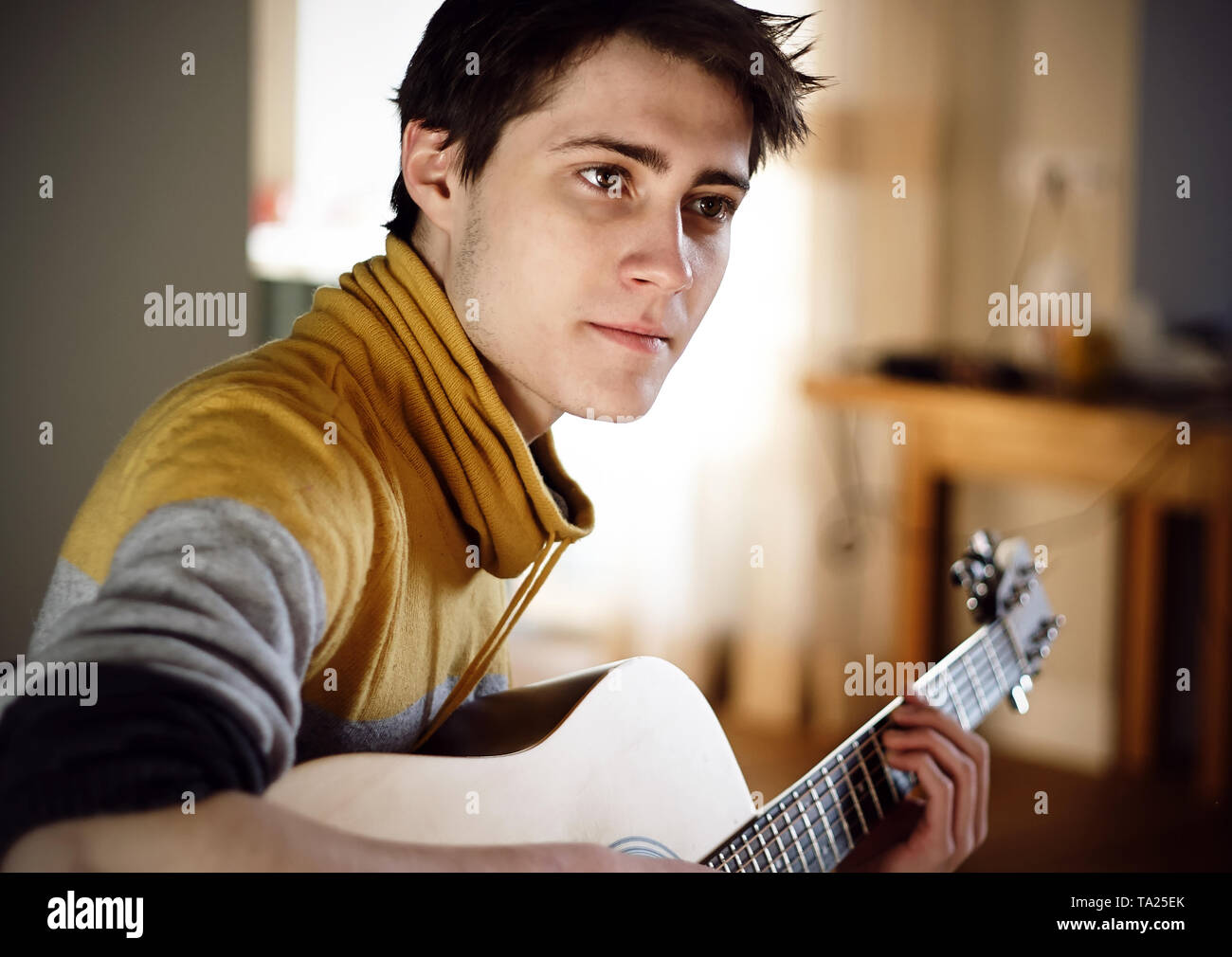 Un beau jeune homme aux yeux bruns avec les cheveux foncés dans un chandail gris-jaune joue une guitare acoustique tout en restant assis à la maison sur une journée ensoleillée. Banque D'Images