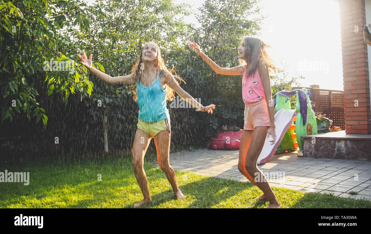 Photo de deux soeurs en rire heureux les vêtements humides danser sous les gouttelettes d'eau d'arrosage au jardin. Le jeu et le plaisir de la famille en plein air au Banque D'Images