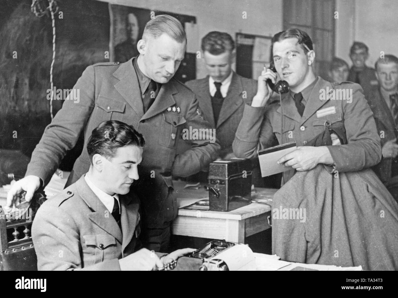 Photo de soldats allemands de la légion Condor mise sur des vêtements civils après la fin de la guerre civile espagnole (avril 1939). Au premier plan il y a un caporal assis près d'une machine à écrire. Derrière lui, un lieutenant est donnant des ordres. Sur la droite, près du téléphone, un caporal avec un crayon derrière l'oreille et vestes sous son bras. Dans l'arrière-plan sont des soldats, qui sont déjà en tenue civile. Banque D'Images