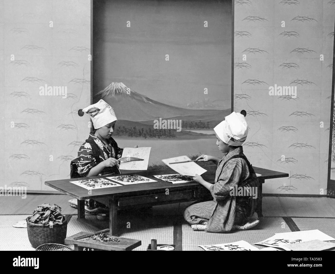 Deux jeunes femmes travaillent avec de l'encre et du papier. Ils sont assis sur le sol à une table basse. Dans l'arrière-plan est une peinture d'une montagne. Banque D'Images