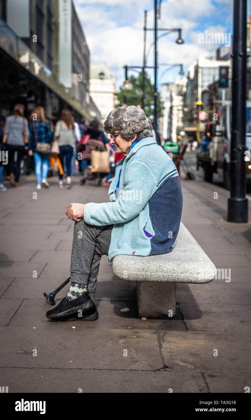 Portrait de vue latérale d'une femme âgée assise seule sur un banc de béton, regardant pensive, Oxford Street, Londres, Angleterre,ROYAUME-UNI. Banque D'Images