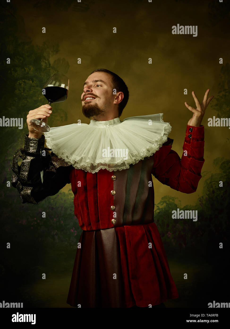 Jeune homme comme un chevalier médiéval sombre sur fond de studio. Portrait de modèle masculin en costume rétro. Tenant un verre de vin rouge. Les émotions humaines, compar Banque D'Images