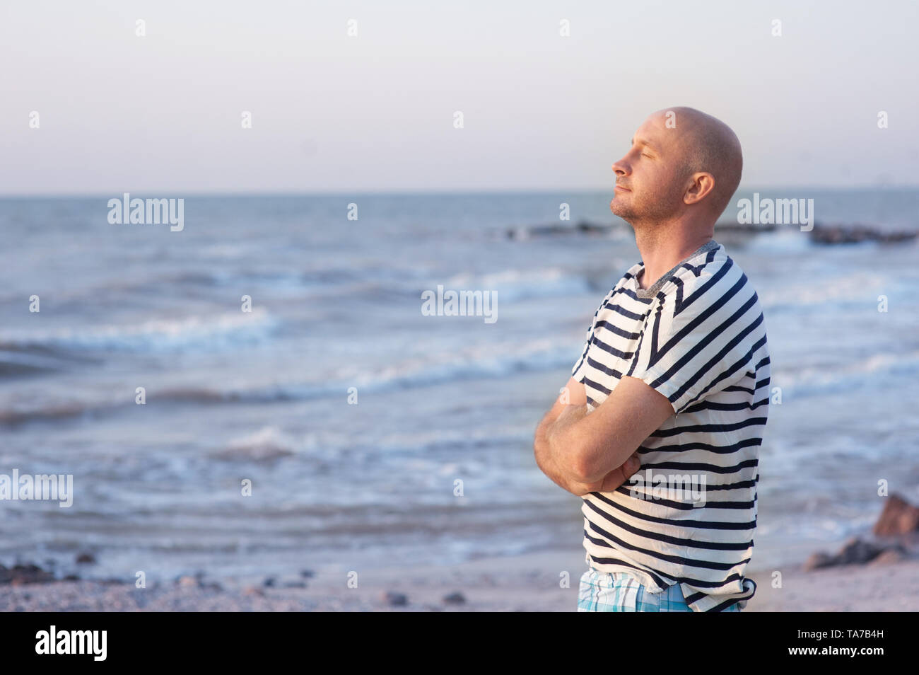 Beau homme portant un t-shirt rayé respirant devant l'océan sur la plage Banque D'Images