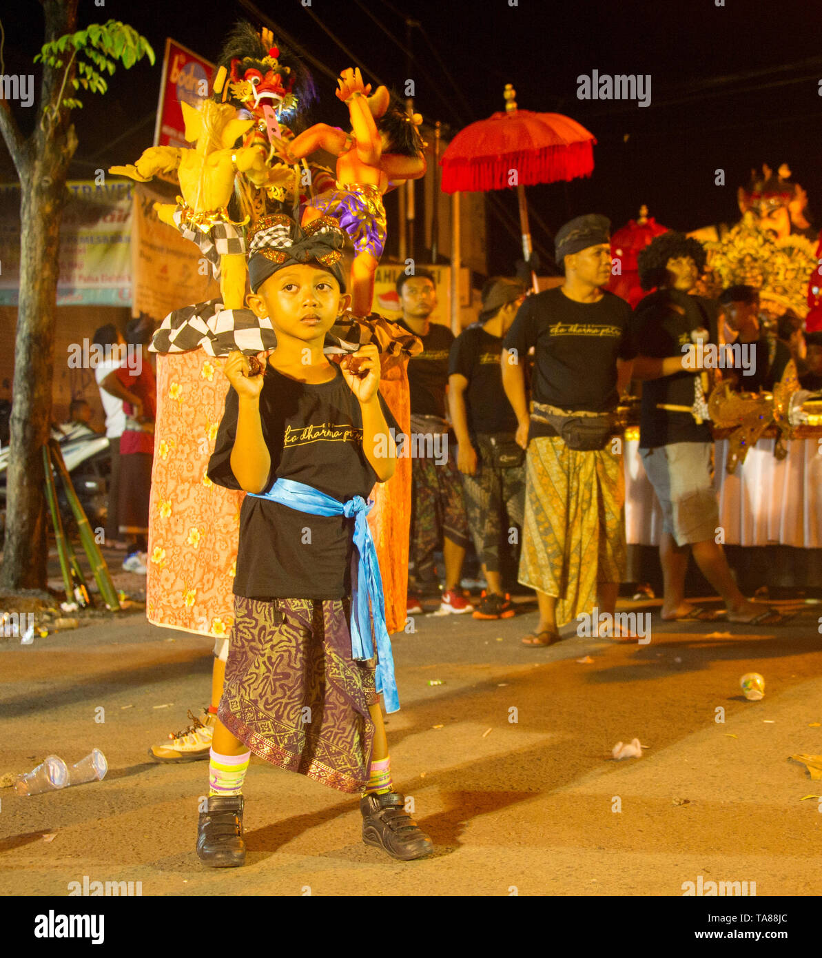 BALI, INDONÉSIE - Mars 27, 2017 : Garçon en costume traditionnel holding chiffres Nyepi, défilé traditionnel dans les rues de Kuta en soirée en arrière-plan Banque D'Images