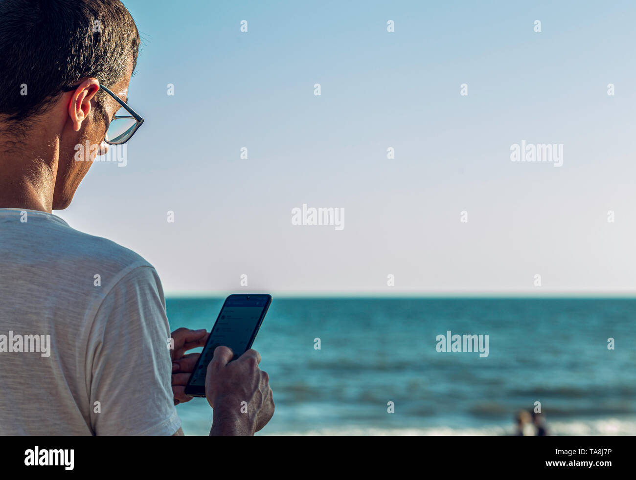 Vue latérale d'un homme séduisant en utilisant un téléphone intelligent sur la plage avec la mer et l'horizon à l'arrière-plan Banque D'Images