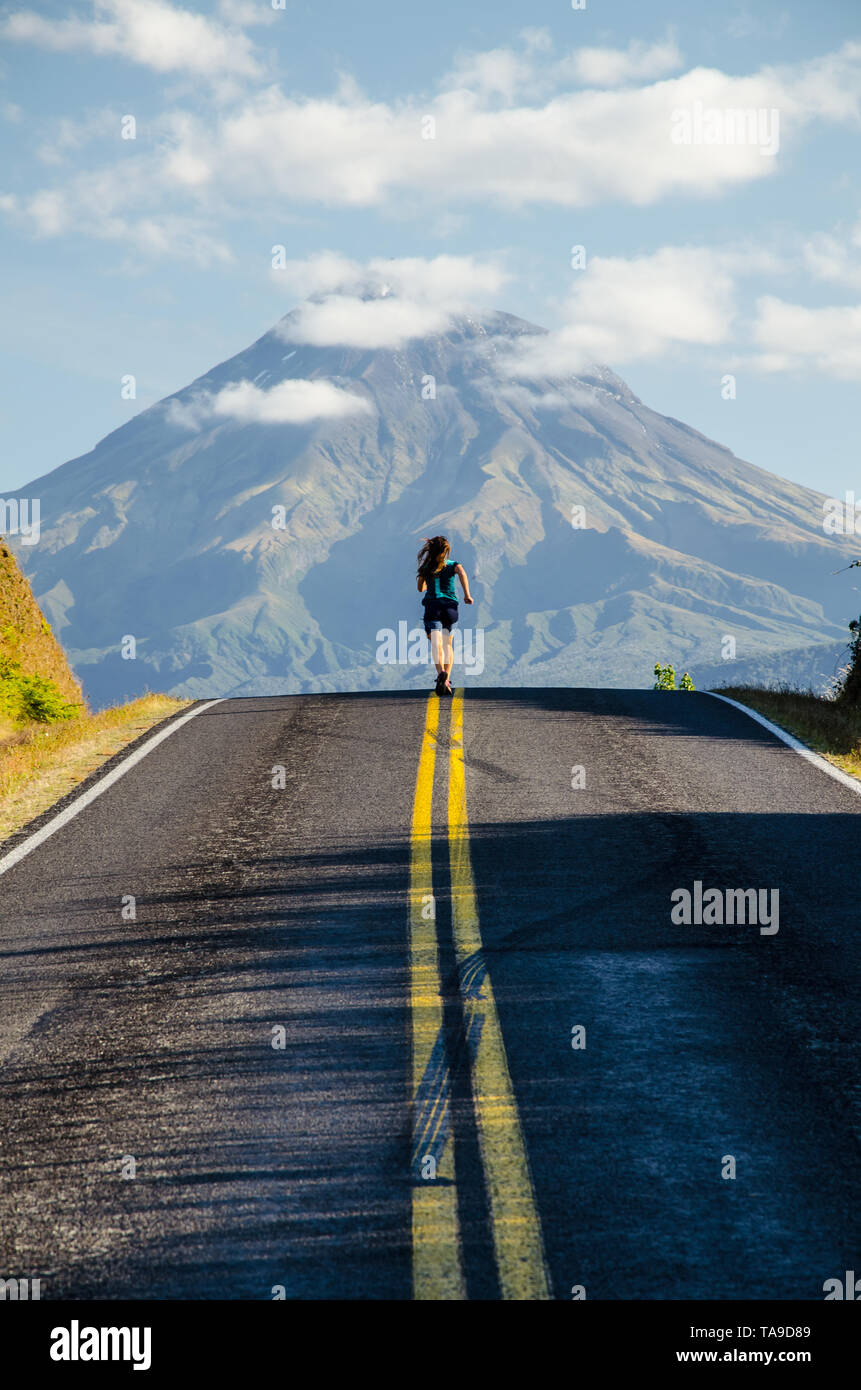 Fille courir en t-shirt vert sur la route de montagne avec en arrière-plan Taranaki avec les nuages au-dessus, Parc National d'Egmont, Nouvelle-Zélande, île du Nord. Banque D'Images