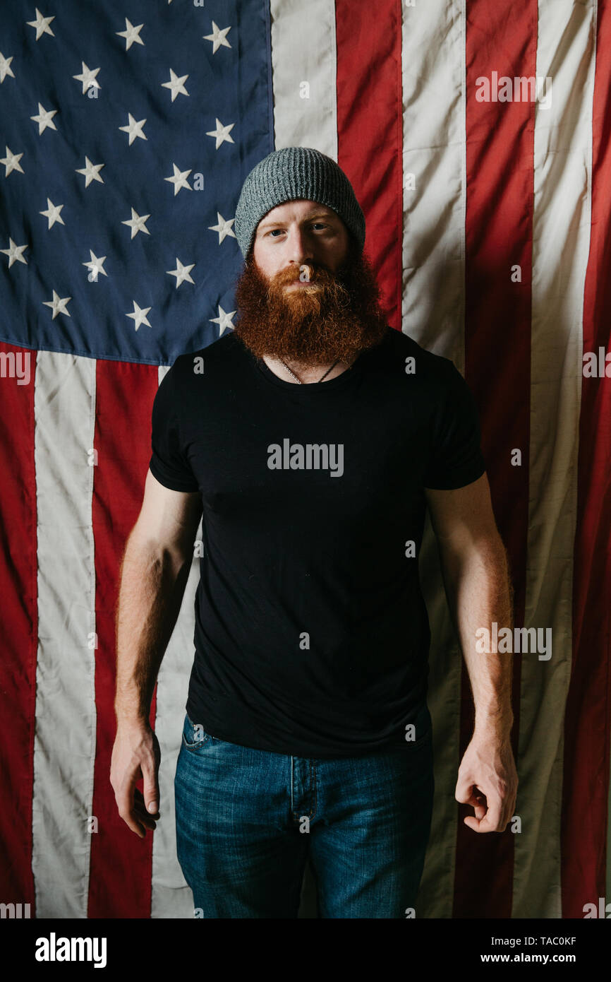 Un jeune homme avec une barbe complète, debout devant un drapeau américain portant un bonnet simple. Banque D'Images