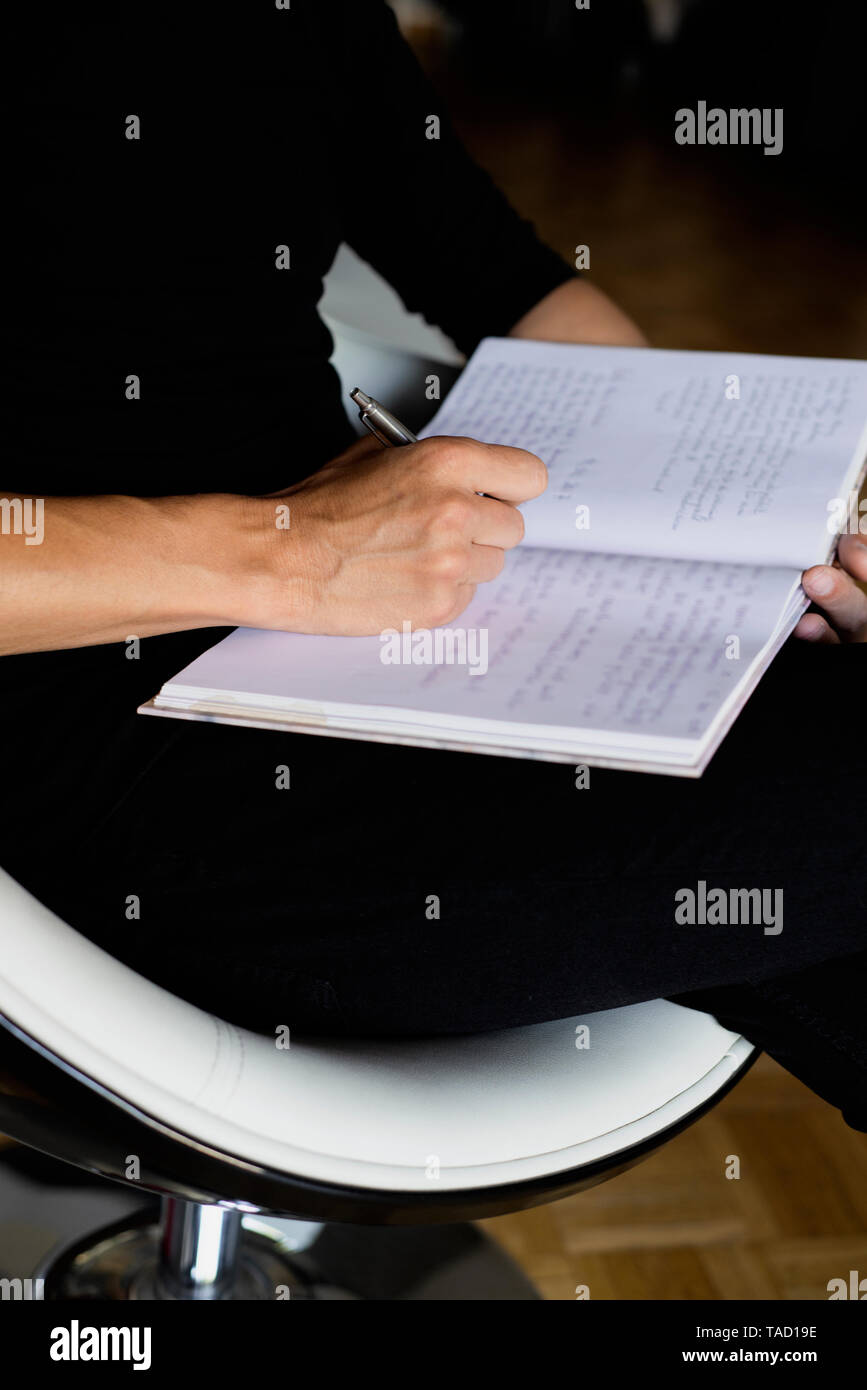 Libre d'un jeune homme de race blanche, portant des vêtements noirs, écrit avec un stylo sur un ordinateur portable assis sur une chaise à l'intérieur Banque D'Images