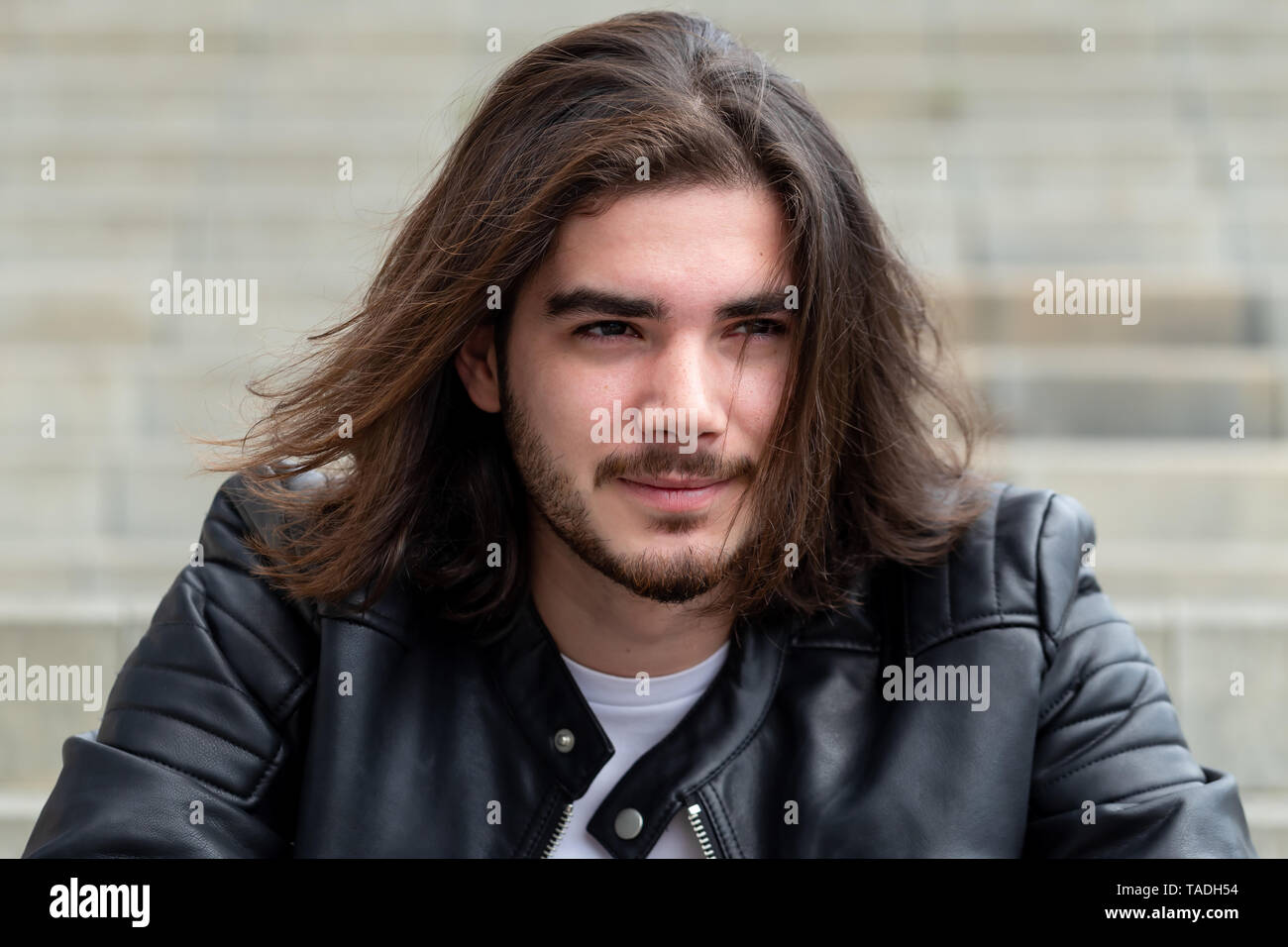 Portrait d'un bel homme barbu avec de longs cheveux bruns, habillé dans les vêtements à la mode, assis sur les marches et à la recherche sur le côté, avec pour zone de contenu publicitaire. Hipster beau mec posant sur l'exemplaire de l'espace. Banque D'Images