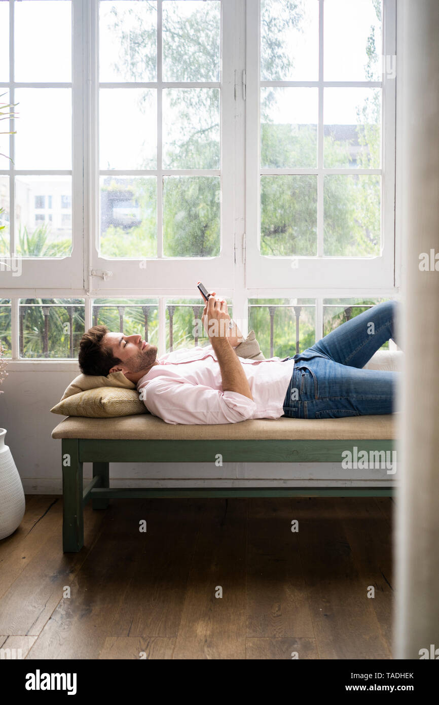 Homme couché sur un banc en face de wondow, using smartphone Banque D'Images