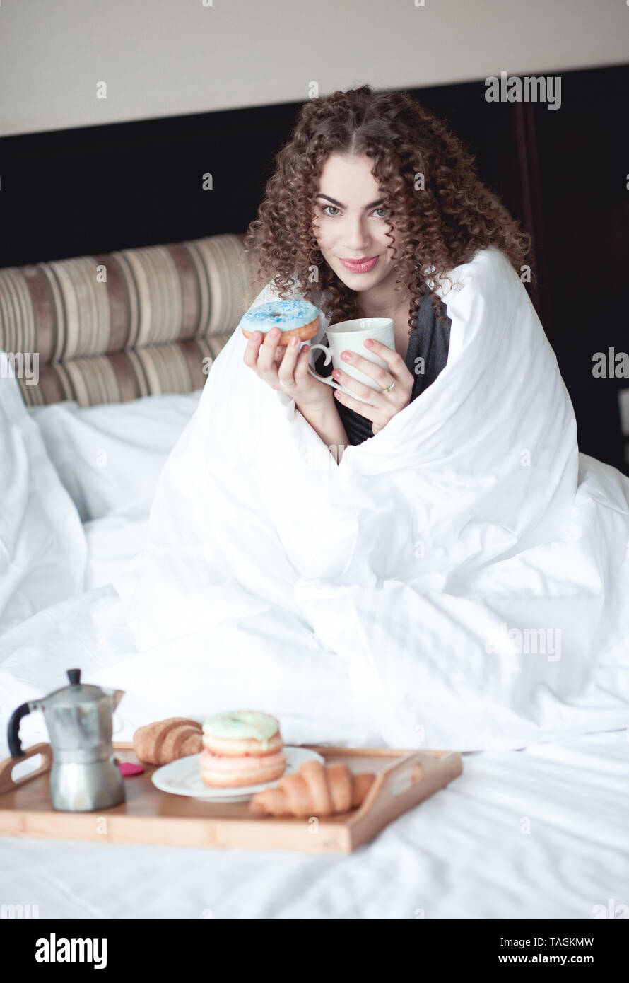 Femme 24 à 29 ans de service au lit de manger des beignes et de boire du café dans la chambre d'hôtel. En regardant la caméra. Bonjour. Banque D'Images