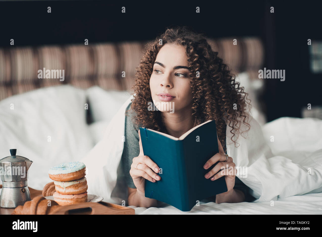 Belle fille 24-29 ans reading book in bed avec petit déjeuner sur plateau en bois dans la chambre d'hôtel. Bonjour. En regardant la caméra. Activité de loisirs. Banque D'Images