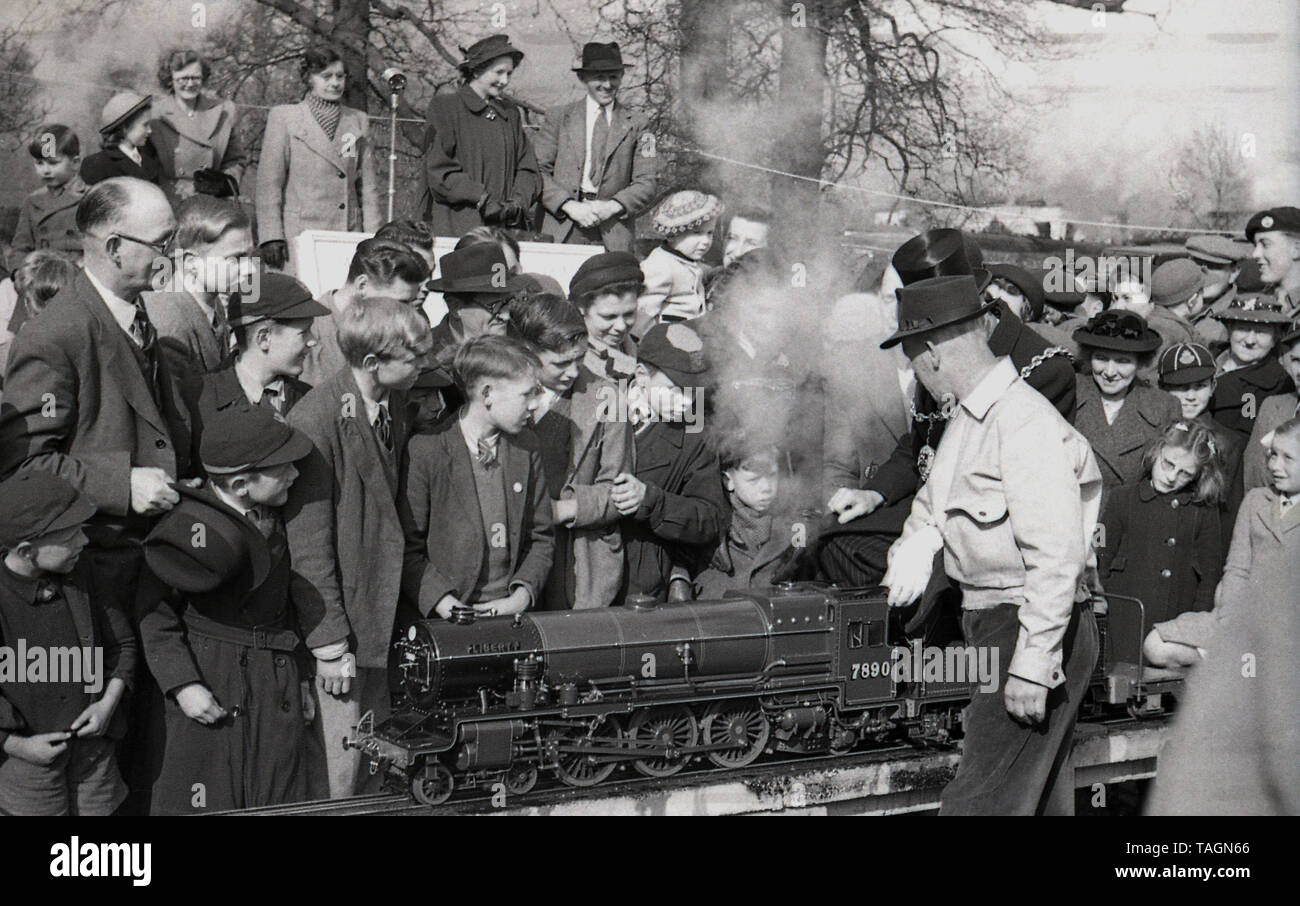Années 1950, historiques, une foule de personnes à la recherche d'un train à vapeur miniature sur un modèle de fer, avec un dignitry possiblly local, le maire ayant un tour sur elle, England, UK. Cet étroit chemin de fer miniature jauge de est relevée au-dessus du sol et le conducteur et les passagers assis à califourchon sur la piste. En cette ère de fer miniature ou modèle avec des locomotives à vapeur ont été une attraction populaire dans les parcs et les villes de bord de mer. Banque D'Images